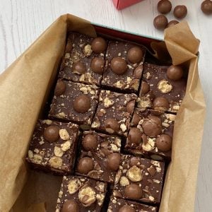 Slices of Malteser Tiffin in a rectangular tin lined with baking parchment. Balls of chocolate maltesers scattered on the side.
